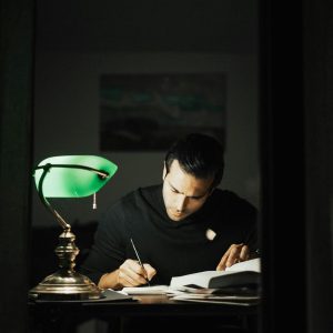 Concentrated young male wearing black turtleneck taking notes and reading books while sitting at desk with papers and bankers lamp in dark home office